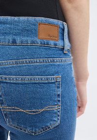 Πίσω όψη ατόμου που φοράει μπλε τζιν παντελόνι με δερμάτινο ταμπελάκι λογότυπου Pepe Jeans πάνω από την πίσω τσέπη, με μαύρο φόντο.