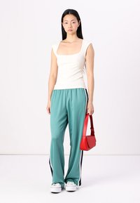 Hvit ribbet tanktopp, teal vid bukse med marineblå stripete sider, og en rød crossover-veske. Modellen står foran en ensfarget bakgrunn.