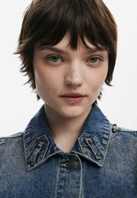 Detailný záber na denimovú bundu s klasickým golierom, tmavomodrou farbou, štruktúrovanou tkaninou a dekoračnými kovovými prvkami na pleciach.