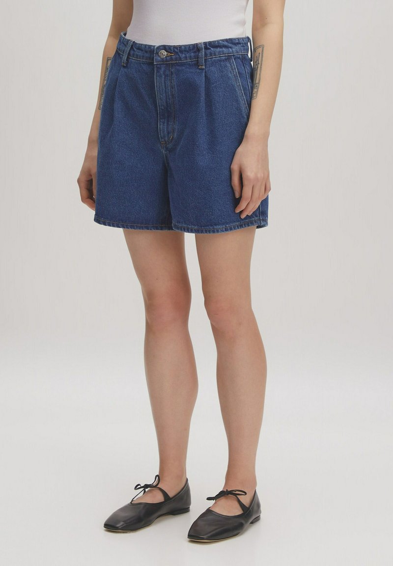 Blaue Denim-Shorts mit hoher Taille, gefälteltem Front und umgeschlagenen Säumen, kombiniert mit schwarzen Ballerinas mit einer Schleifenverzierung.