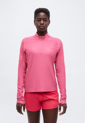 Femme noire debout, face à l'avant, portant un haut de sport rose à manches longues avec fermeture éclair et un short rose assorti sur un fond uni.