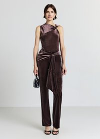 Combinaison en velours violet foncé avec un décolleté asymétrique et un drapé à la taille. Associée à un petit sac à main noir et des talons.