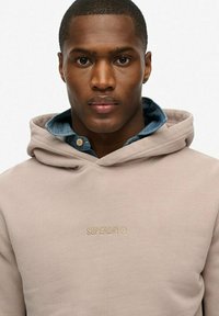 Beige hooded sweatshirt with a front logo, made of a soft fabric, layered over a blue denim shirt, featuring a relaxed fit and smooth texture.