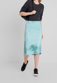 En midjelang satängskjol i en ljus turkos färg, med en slät yta och en lätt lyster, i kombination med en mörk tröja och svarta sneakers.