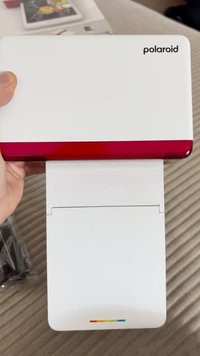 Imprimante Polaroid blanche avec un dessus rouge brillant. L'avant présente une fente pour le papier et une petite bande multicolore en bas.