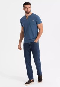 Camisa henley de manga corta azul con un bolsillo frontal, combinada con jeans de mezclilla oscura y calzado marrón. Diseño simple, ajuste relajado.