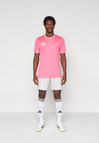 Ροζ αθλητική φανέλα με λευκή επένδυση, συνδυασμένη με γκρι σορτς, μαύρες και λευκές ριγωτές κάλτσες και ποδοσφαιρικά παπούτσια. Λογότυπο Adidas ορατό.