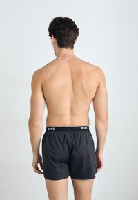 Boxers noirs avec une taille élastique affichant "BOSS" en blanc, fabriqués à partir d'un tissu lisse et léger. Vu de l'arrière.