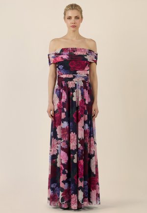 Frau in bodenlangem, schulterfreiem Kleid vor dunklem Hintergrund mit großem pink-, lila- und rotfarbenem Blumenmuster.