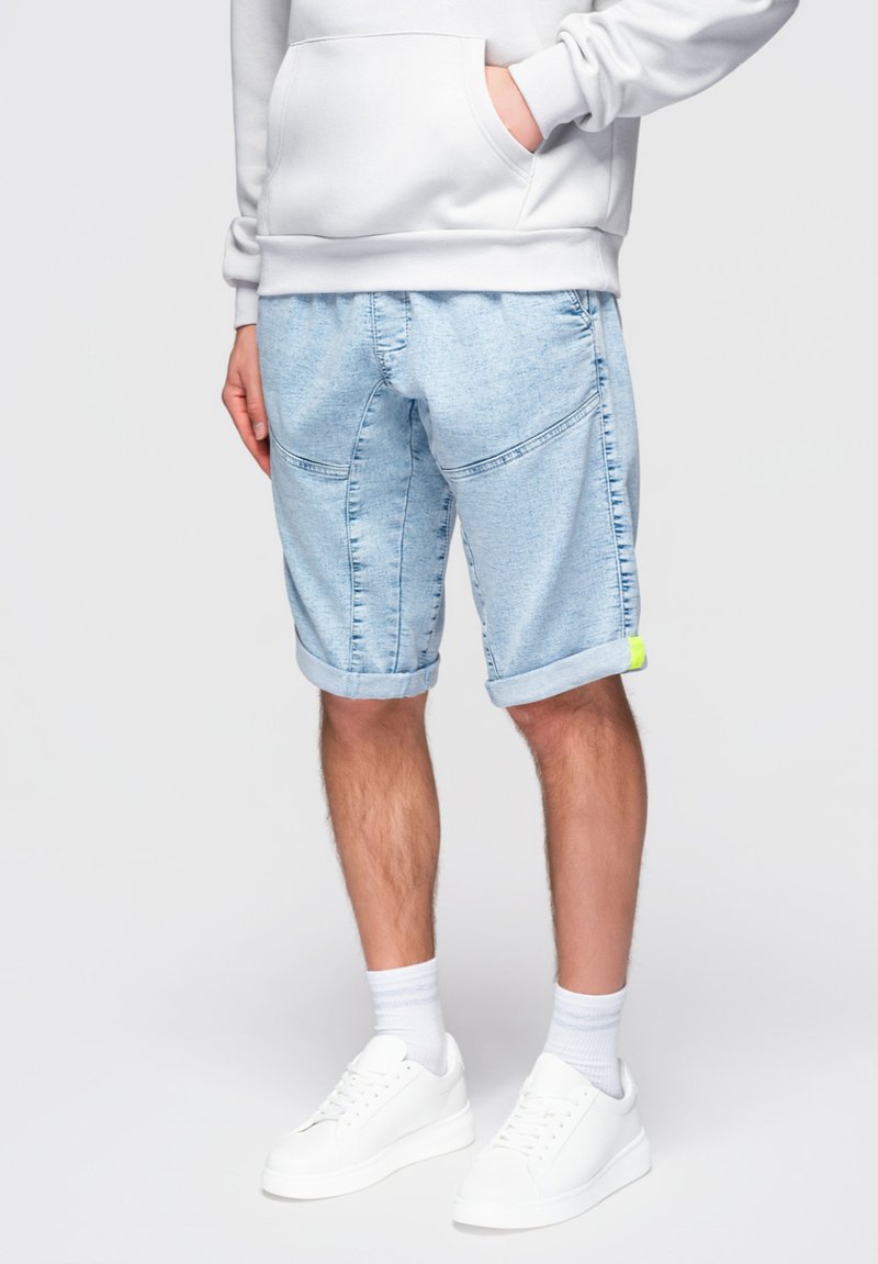 Person iført lyseblå denimshorts med op-rullede kanter og en hvid sweatshirt, stående med den ene hånd i lommen.
