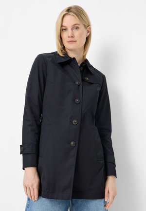 Blonde woman wearing a black buttoned coat with long sleeves and blue jeans, standing against a white background.