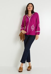Cardigan en tricot fuchsia avec broderies florales, capuche, franges, associé à un jean en denim foncé et des mules à talons carrés en daim marron.