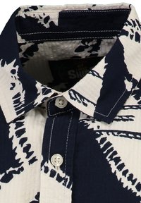 Kurzarm-Hemd mit Knopfleiste in Creme und Marineblau, mit einem Palmblattmuster, aus einem strukturierten Stoff und mit einem spitzen Kragen. Weiße Knöpfe sichtbar.