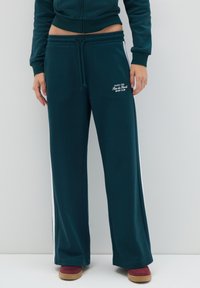 Mujer vistiendo pantalones de chándal de pierna ancha en verde oscuro con franjas laterales blancas y un logo blanco del Club Deportivo Paris 1996 Rue de Rivoli.