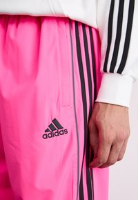 Adidas pink track pants feature black side stripes, a smooth texture, and an embroidered logo near the pocket. Contrast cuffs complete the design.