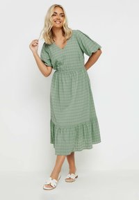 Vihreä ruudullinen midimekko, jossa on V-pääntie, lyhyet puffihihat ja röyhelöhelma. Yhdistetty valkoisiin tasapohjaisiin sandaaleihin.