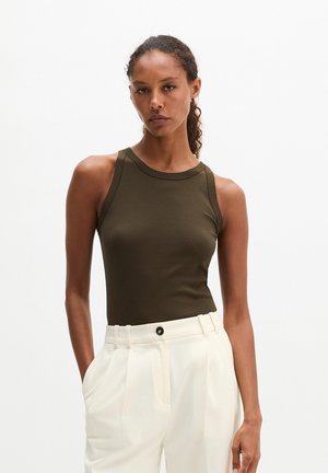 Femme portant un haut sans manches vert olive et un pantalon plissé blanc taille haute, les mains dans les poches, sur un fond uni.