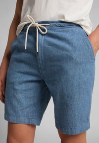 Pantalones cortos de mezclilla azul con cintura ajustable, dos bolsillos y una textura tejida. El diseño incluye un dobladillo con vuelta y detalles de costura sutiles.