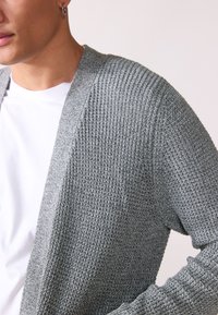 Grå stickad cardigan med en texturerad mönster, öppen front och långa ärmar, bärs över en vit t-shirt.