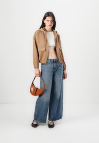 Veste à capuche zippée marron, crop top blanc, jeans en denim à jambes larges, chaussures noires à lacets et un petit sac à main marron. Texture lisse, design décontracté.
