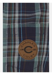 Karierte Flanellbluse in Navy-, Teal- und Brauntönen mit einem Leder-Patch, das die Marke "Charles Colby" auf der linken Brusttasche zeigt.