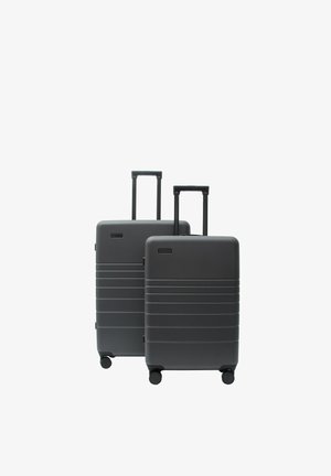 Dwie twarde walizki w ciemnoszarym kolorze z wysuwanymi uchwytami i czterema kółkami, jedna większa i jedna mniejsza, stojące obok siebie na białym tle.
