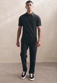 Polo shirt negru cu mâneci scurte, combinat cu pantaloni întunecați și adidași negri cu șireturi albe, stând pe un fundal neutru.