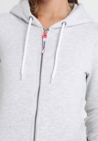 Sudadera con cremallera de color gris claro, con capucha ajustable y tirador de cremallera rosa, además de cuerdas de acento blancas. Textura suave y diseño casual.