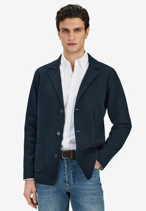 Junger Mann trägt dunkelblauen Blazer über weißem Hemd und blauer Jeans, steht mit einer Hand in der Tasche vor weißem Hintergrund.