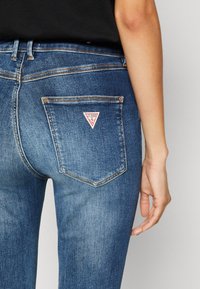 Tamno plave traperice imaju prilagođen dizajn s trokutastim zakrpun na stražnjem džepu koji prikazuje "GUESS" logo i vidljivim šivom.