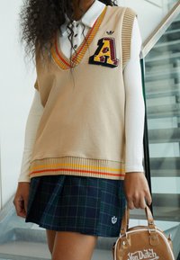Beige stickad väst med orange och gulrandig kant och ett bokstavspatch. Bärs över en vit långärmad skjorta och en rutig marinblå kjol.