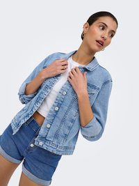 Woman wearing light blue denim jacket over white top and dark blue denim shorts, holding jacket collar and looking to the side.