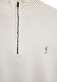 Kremowy dzianinowy sweter z krótkim czarnym zamkiem i małym haftowanym logo przedstawiającym osobę grającą na rogu na prawej piersi.