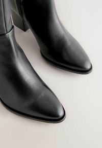 Bottines en cuir noir avec un bout pointu et un talon moyen en bloc. Texture lisse et détail de fermeture éclair sur le côté pour un enfilage facile.
