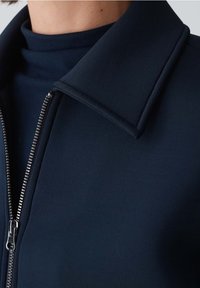 Giacca zip blu navy con colletto alto, realizzata in un tessuto morbido ed elastico. Presenta una zip argentata e un design elegante e minimalista.