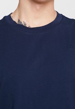 Cou masculin avec un t-shirt bleu marine à col rond arborant un logo discret en relief sur le côté gauche, avec un petit tatouage visible sur le cou.
