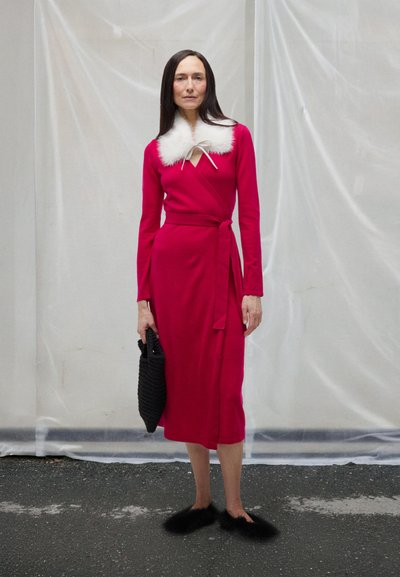 Nő, aki hosszú piros, fehér szőrmével díszített vállkendőruhát visel, kezében fekete fonott táskával, járdán áll, világos szövet háttér előtt.