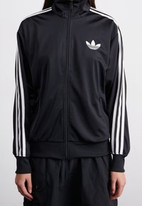 Chaqueta negra con cremallera, hecha de un material suave, con rayas blancas a lo largo de las mangas y un pequeño logo blanco en la parte superior izquierda del pecho.