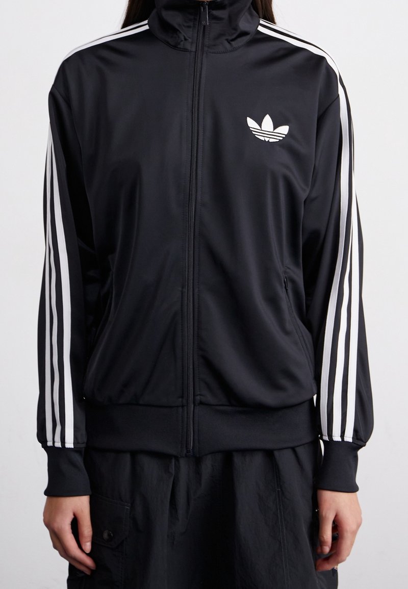 Chaqueta negra con cremallera, hecha de un material suave, con rayas blancas a lo largo de las mangas y un pequeño logo blanco en la parte superior izquierda del pecho.