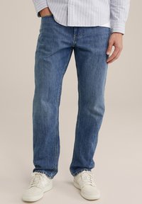 Blauwe denimjeans met een rechte pasvorm, voorzien van subtiele vervaging en stikseldetails. Gecombineerd met witte sneakers, gepresenteerd op een neutrale achtergrond.