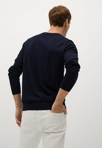 Maglione lavorato a maglia blu navy con maniche lunghe, polsini e orlo a coste, indossato sopra pantaloni bianchi. La vista dal retro mostra una texture morbida e un design semplice.