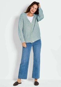 Helles blaues Strickcardigan mit V-Ausschnitt, Knopfverschluss, gerippten Bündchen und lockerer Passform; kombiniert mit blauen Jeans und strukturierten Slippern.