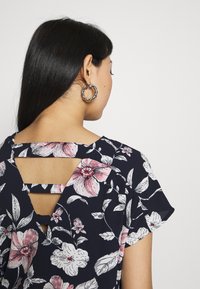 Haut à imprimé floral bleu marine, avec manches courtes et une découpe unique dans le dos. Le tissu est léger et texturé, orné d'accents de fleurs roses.