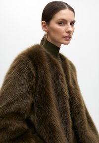 Brown faux fur coat with a soft, dense texture. High collar design and a fitted silhouette. Worn over a dark green turtleneck. Gold hoop earrings.