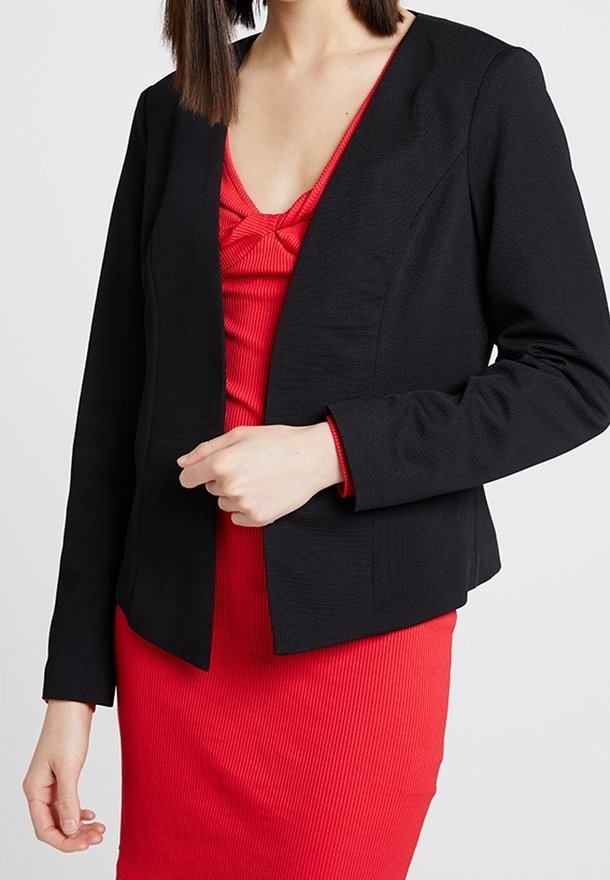 Femme portant un blazer noir ajusté sur une robe rouge côtelée avec un col noué, debout devant un fond clair uni.
