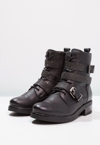 Botas de tobillo de cuero negro con superficie texturizada, múltiples correas decorativas, acentos de tachuelas y una cremallera lateral; tacón bajo tipo bloque.
