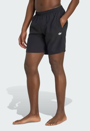 Persona che indossa pantaloncini neri Adidas, in piedi scalza con una mano in tasca su sfondo uniforme.