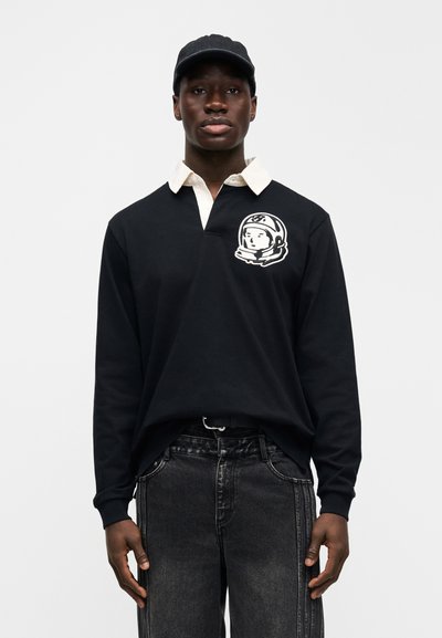 Μαύρο πόλο μακρύ μανίκι με κρεμ γιακά και γραφικό αστροναύτη στο στήθος, συνδυασμένο με σκούρο τζιν παντελόνι. Υλικό από λείο βαμβάκι.