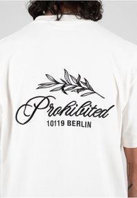 Hvit t-skjorte laget av bomull med et svart brodert design som har teksten "Forbudt" og "10119 BERLIN" sammen med et bladmotiv.