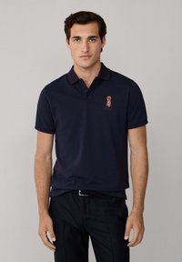 Mörkblå pikétröja med ribbad krage, korta ärmar och ett broderat emblem på vänster bröst. Slät textur och normal passform.